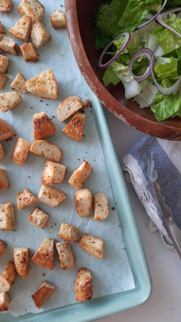 partial view of toasted bagel croutons on a baking sheet and a bowl of cesar salad