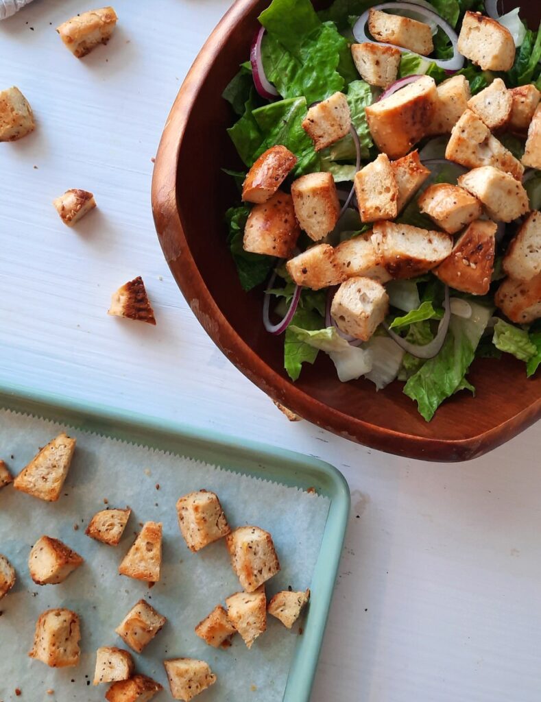 a bowl full of salad covered in bagel croutons sits on a white counter