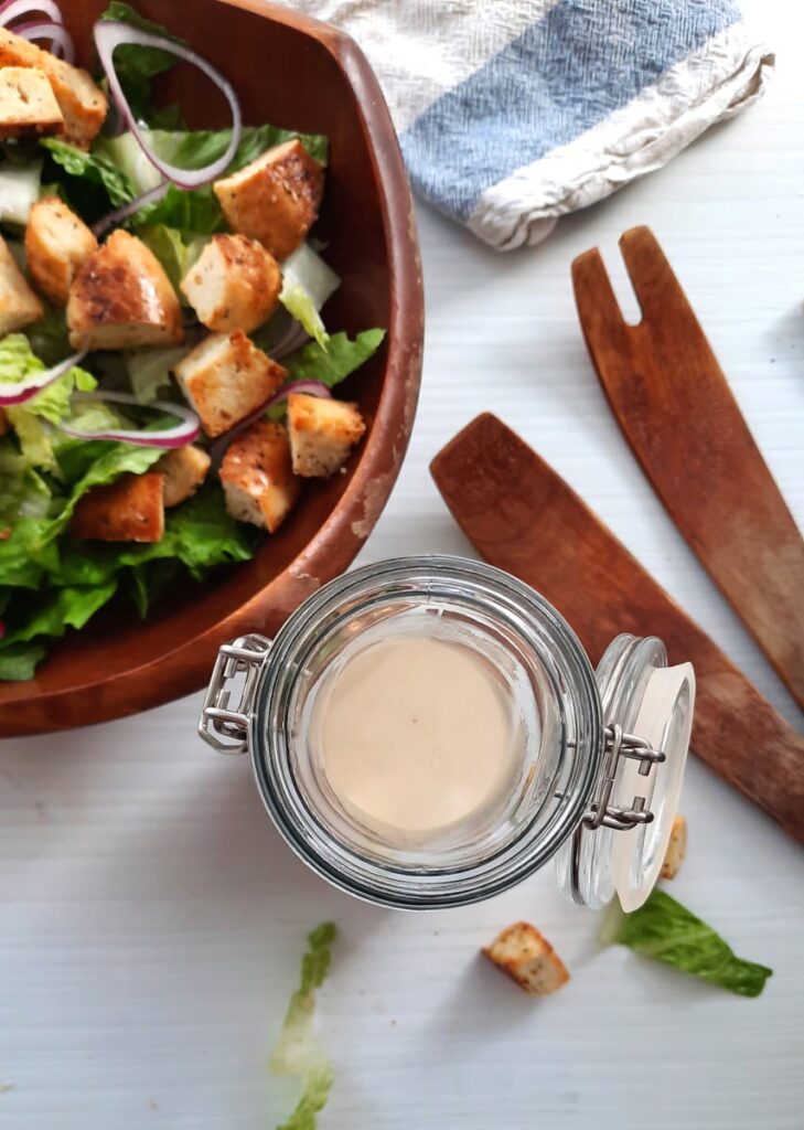 a glass jar full of homemade caesar dressing sits on a couter beside a bowl of caesar salad