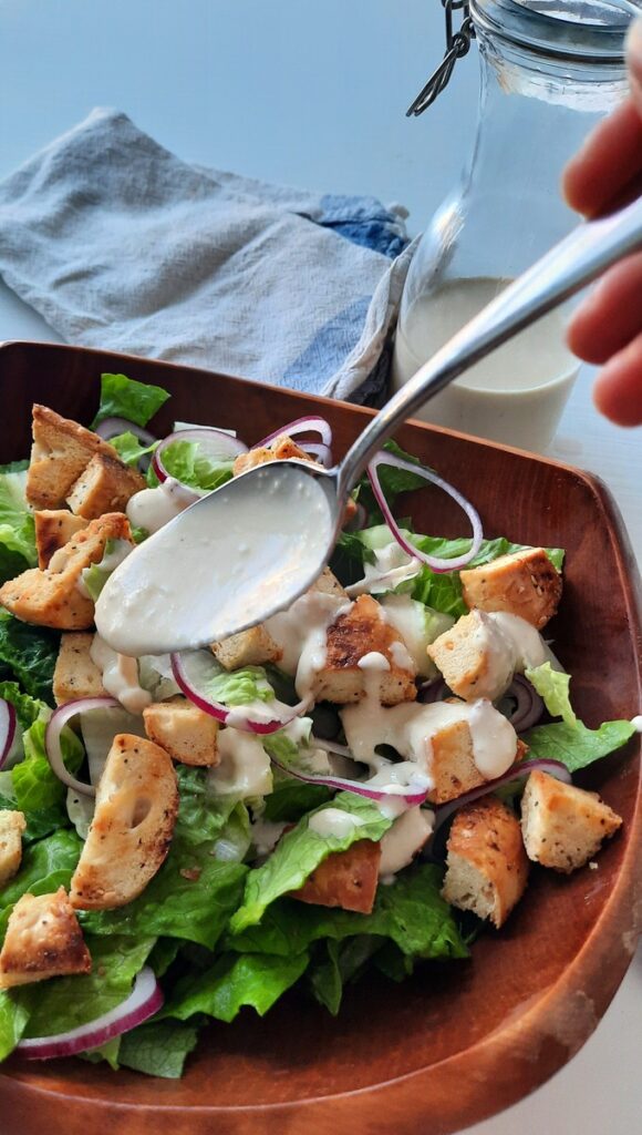 a hand spoons creamy caesar salad dressing all over a salad
