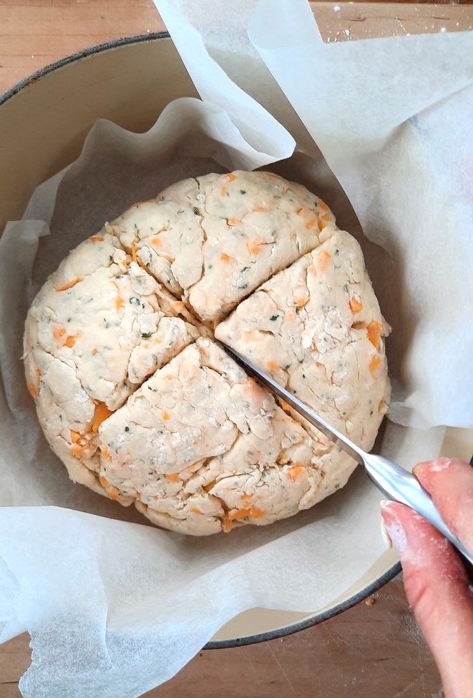 a hand slices an x into soda bread dough shaped into a ball in a dutch oven