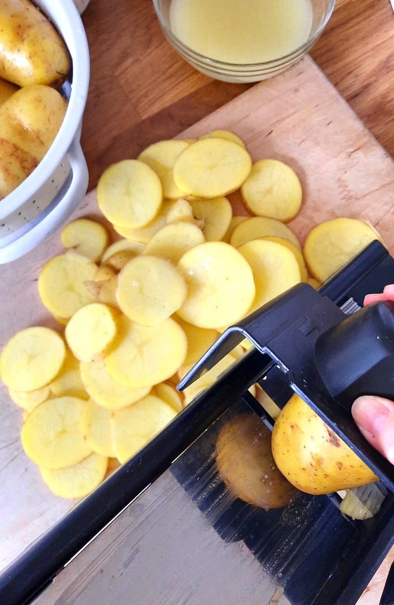 a hand holds a potato in a mandoline to make even slices of potato