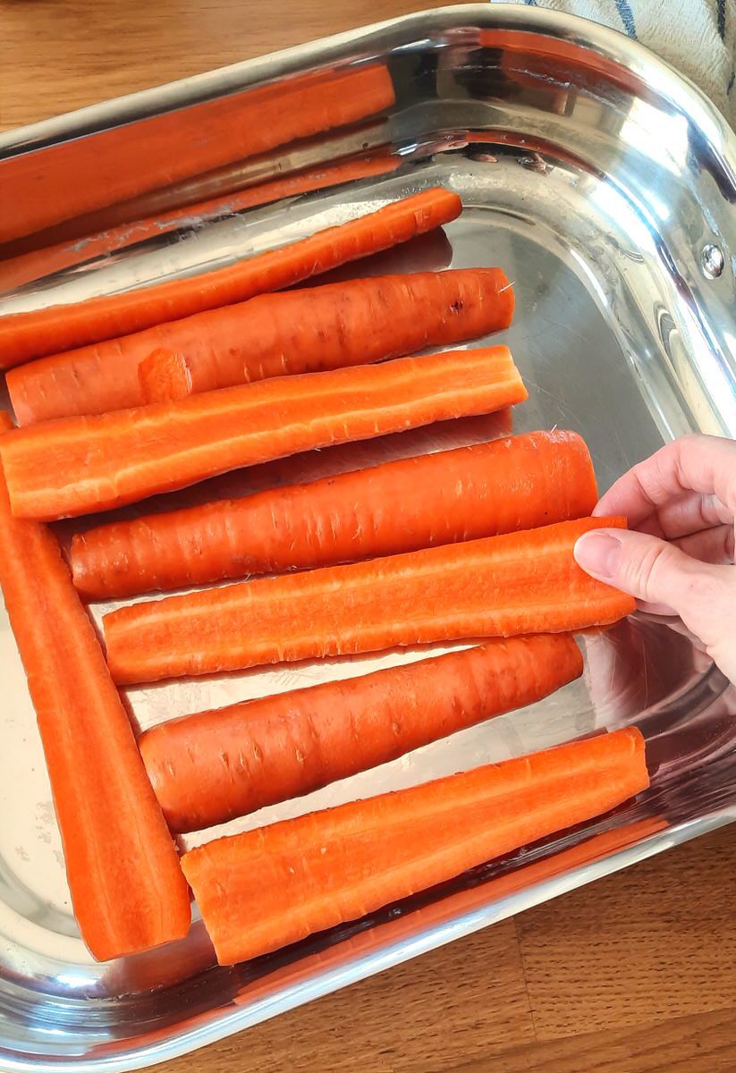 a hand is placing large, halved carrots in the bottom of a roasting pan