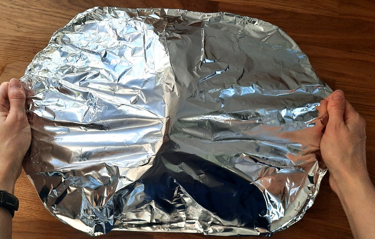 two hands hold a large pan with tinfoil covering a ham inside it