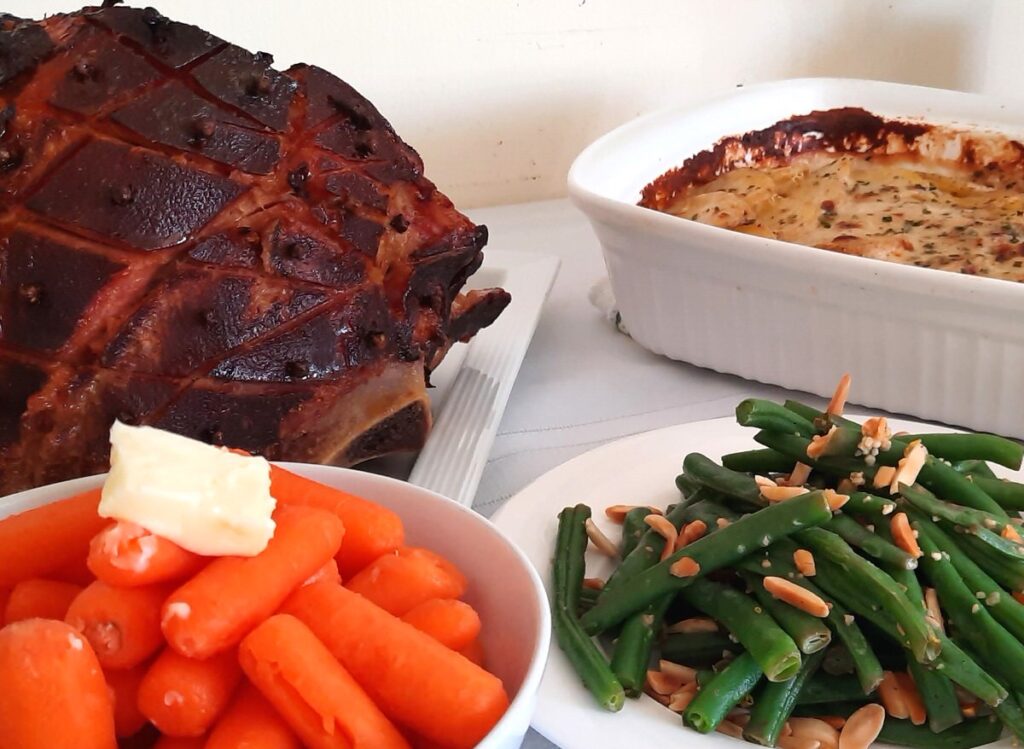 a table with a white tablecloth is covered with steamed carrots, green beans, scalloped potatoes, and a baked ham