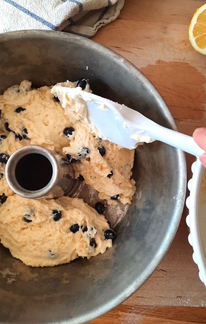 a rubber spatula spoons coffee cake batter with blueberries into a tube pan