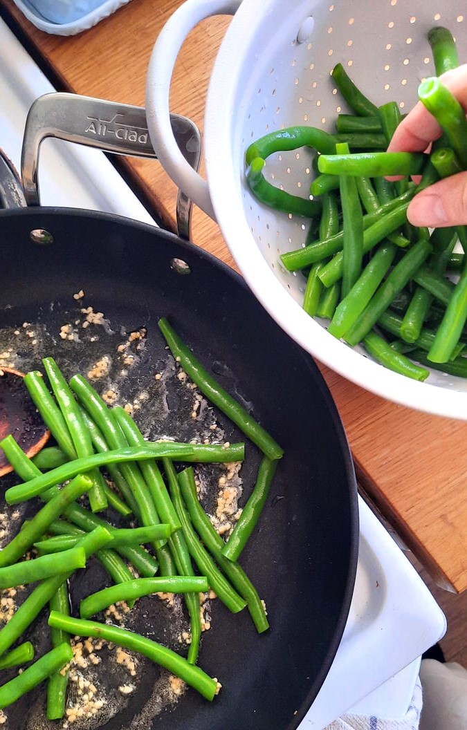 a hand transfers blanched green beans into a hot skillet