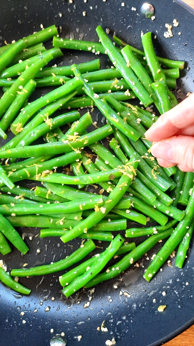 a hand sprinkles lemon zest over a pan of green beans