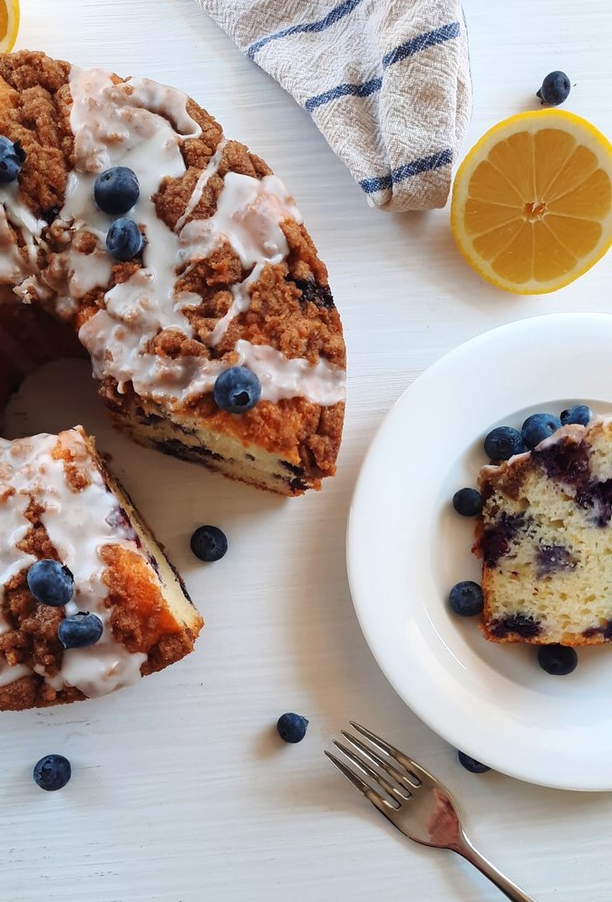 a slice of lemon blueberry coffee cake sits on a plate beside an entire cake with a piece missing