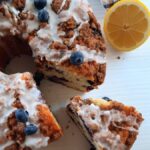 a slice of lemon blueberry coffee cake with a slice removed and resting beside it surrounded by lemons and fresh blueberries