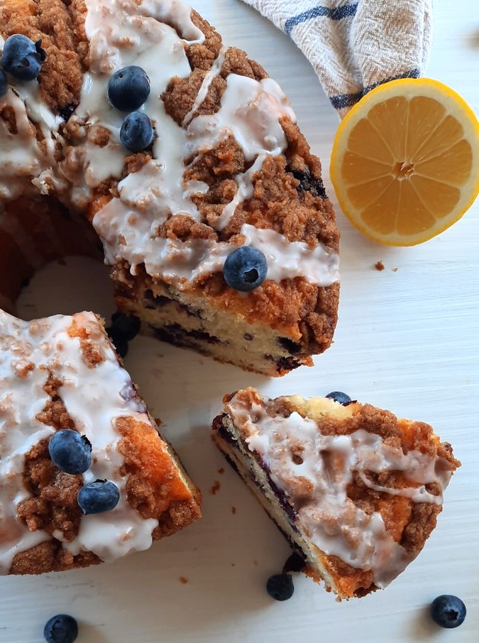 a slice of lemon blueberry coffee cake with a slice removed and resting beside it surrounded by lemons and fresh blueberries
