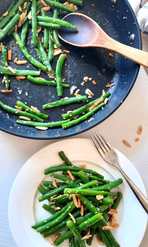a pile of green beans with garlic and lemon and almonds sit on a plate beside a pan with the remaining beans in it