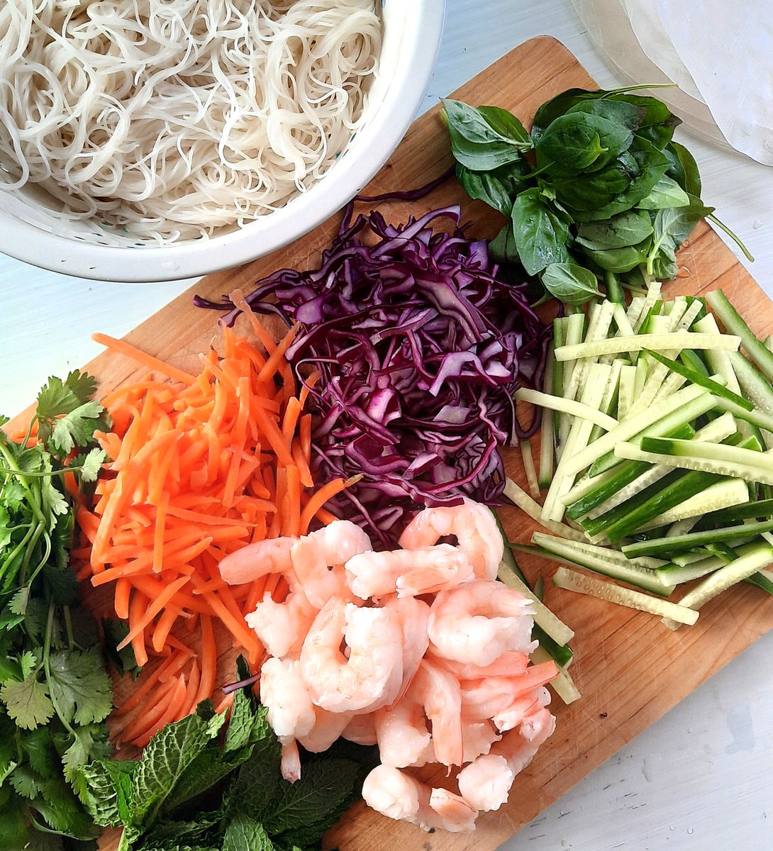 fresh veggies and shrimp on a cutting board beside a bowl of cooked vermicielli noodles and rice paper wrappers