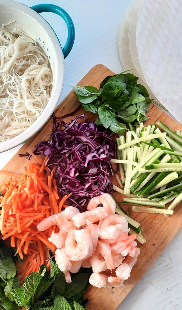 a cutting board filled with fresh spring rolls ingredients like carrot, red cabbage, shrimp, cucumber, and herbs