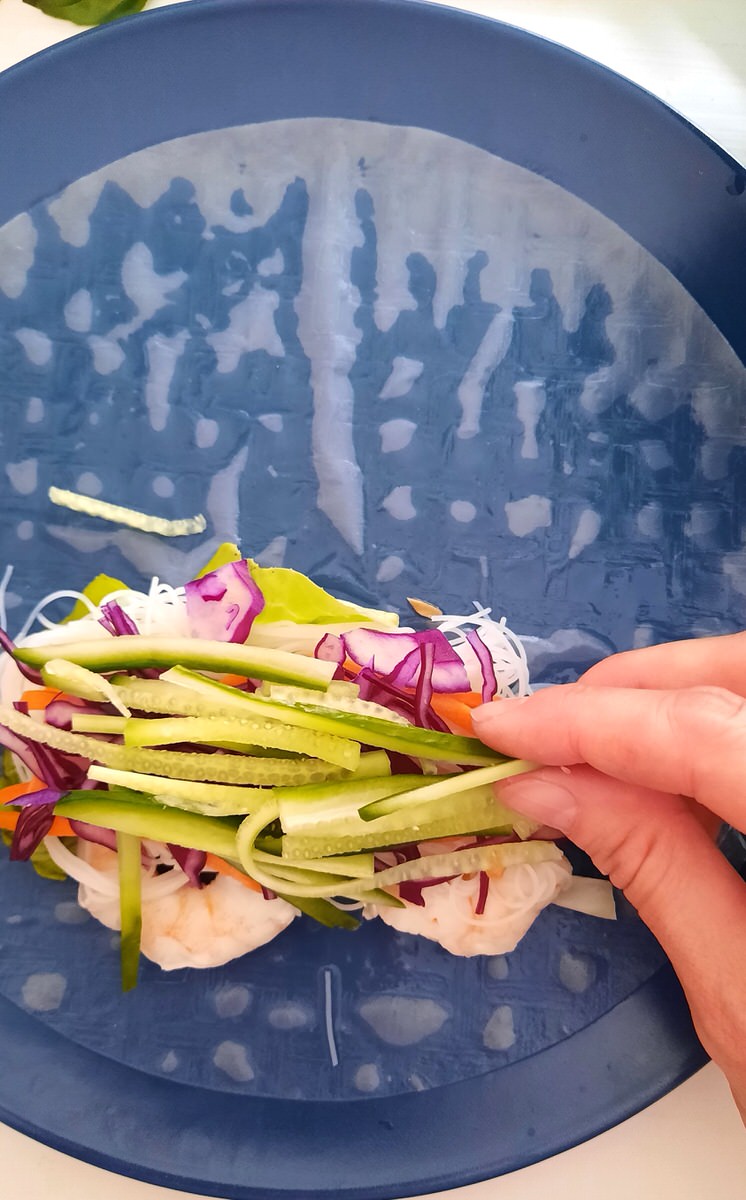 a hand lays julienned cucumber onto a fresh spring roll filled with other veggies, shrimp, and noodles