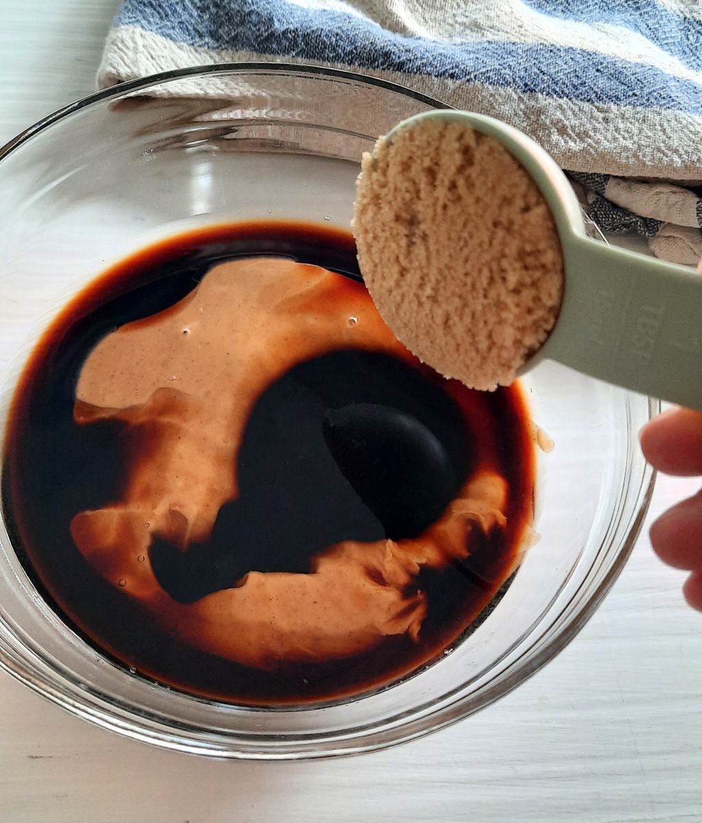 pouring a tablespoon of brown sugar in to a bowl with peanut butter and soya sauce in it to make peanut sauce