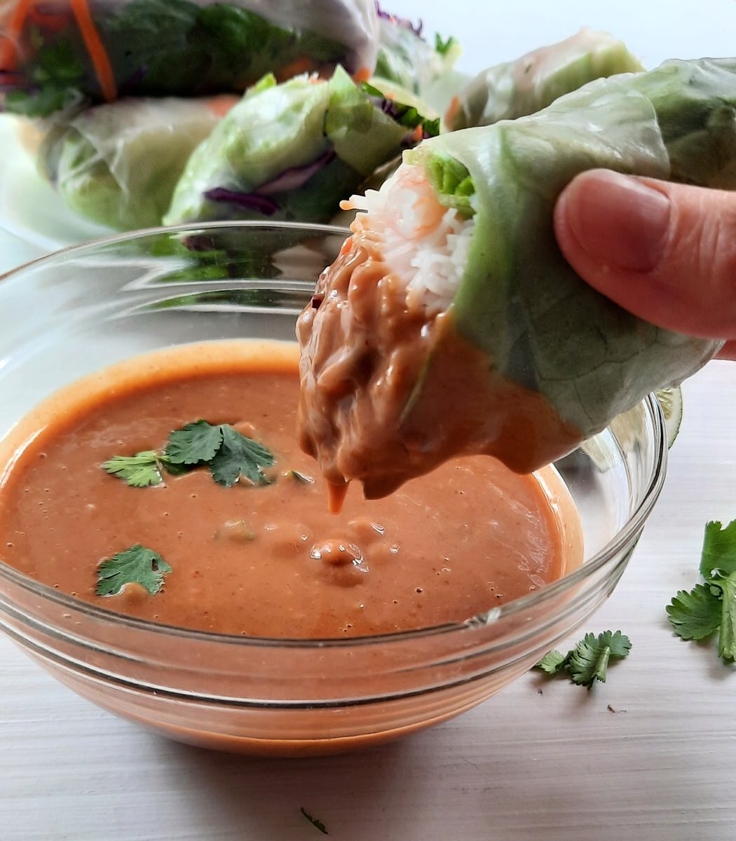 a hand dips half of a fresh spring roll into a bowl of peanut dipping sauce