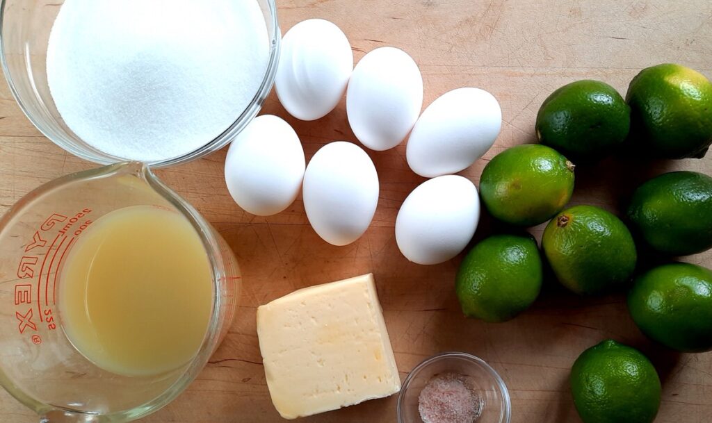 butter, lime juice, eggs, sugar, and limes sit on a cutting board for making lime curd